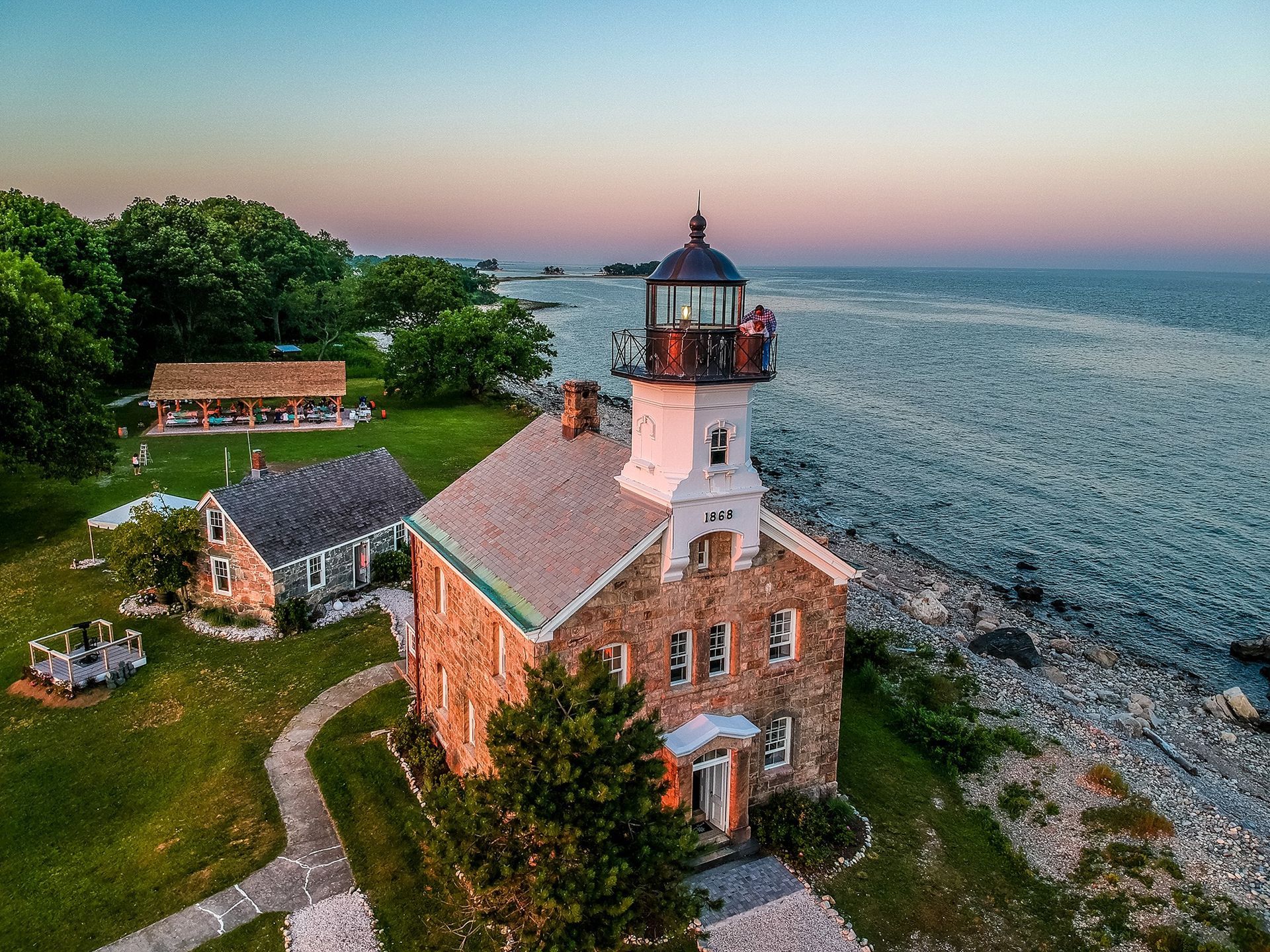 Lighthouse on rocky shore at dusk with green lawn, trees, and small buildings.