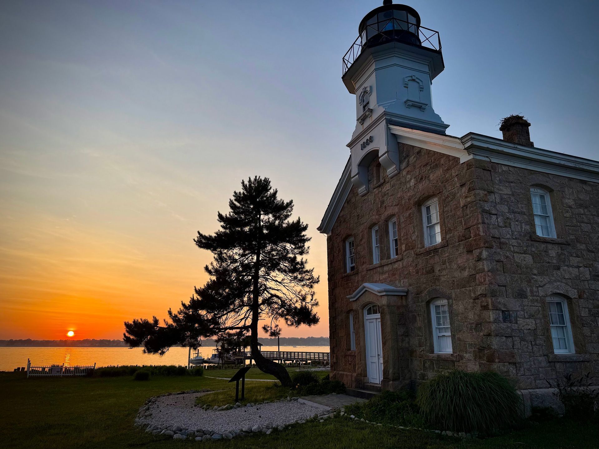 Lighthouse at sunset, stone building with attached tower. Orange and yellow sky over water. Tree silhouette.