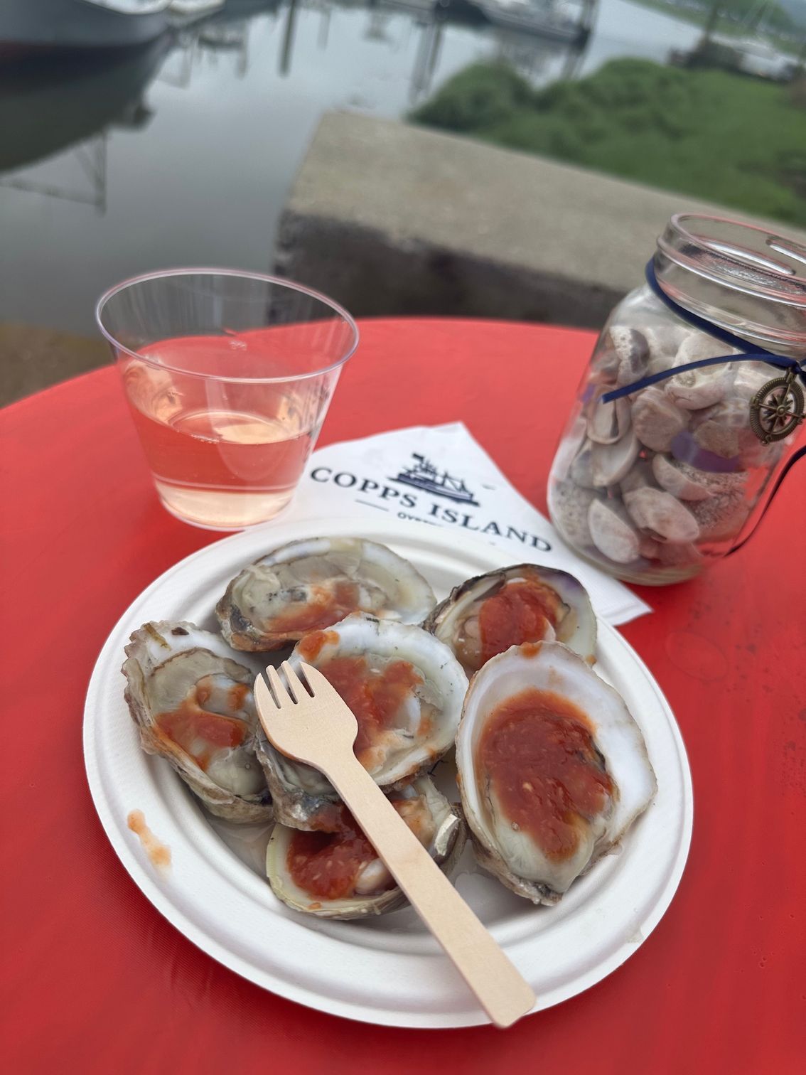 Seafood oysters on a plate with a plastic fork, next to a drink and a jar of sea urchin on a table by water