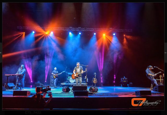 A band performing on stage, bathed in blue and orange lights, with a photographer in the foreground.