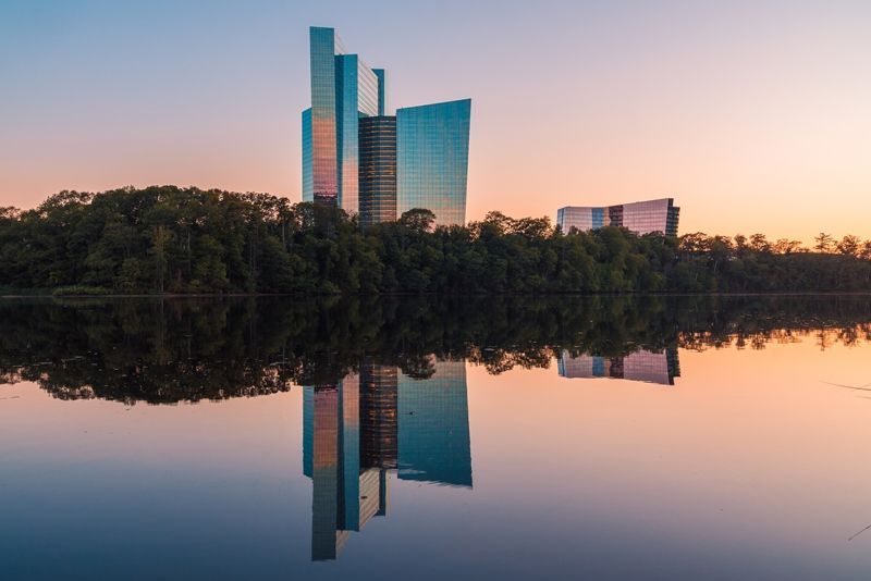 Glass skyscraper reflecting in calm water at dawn, set against a tree line and colorful sky.