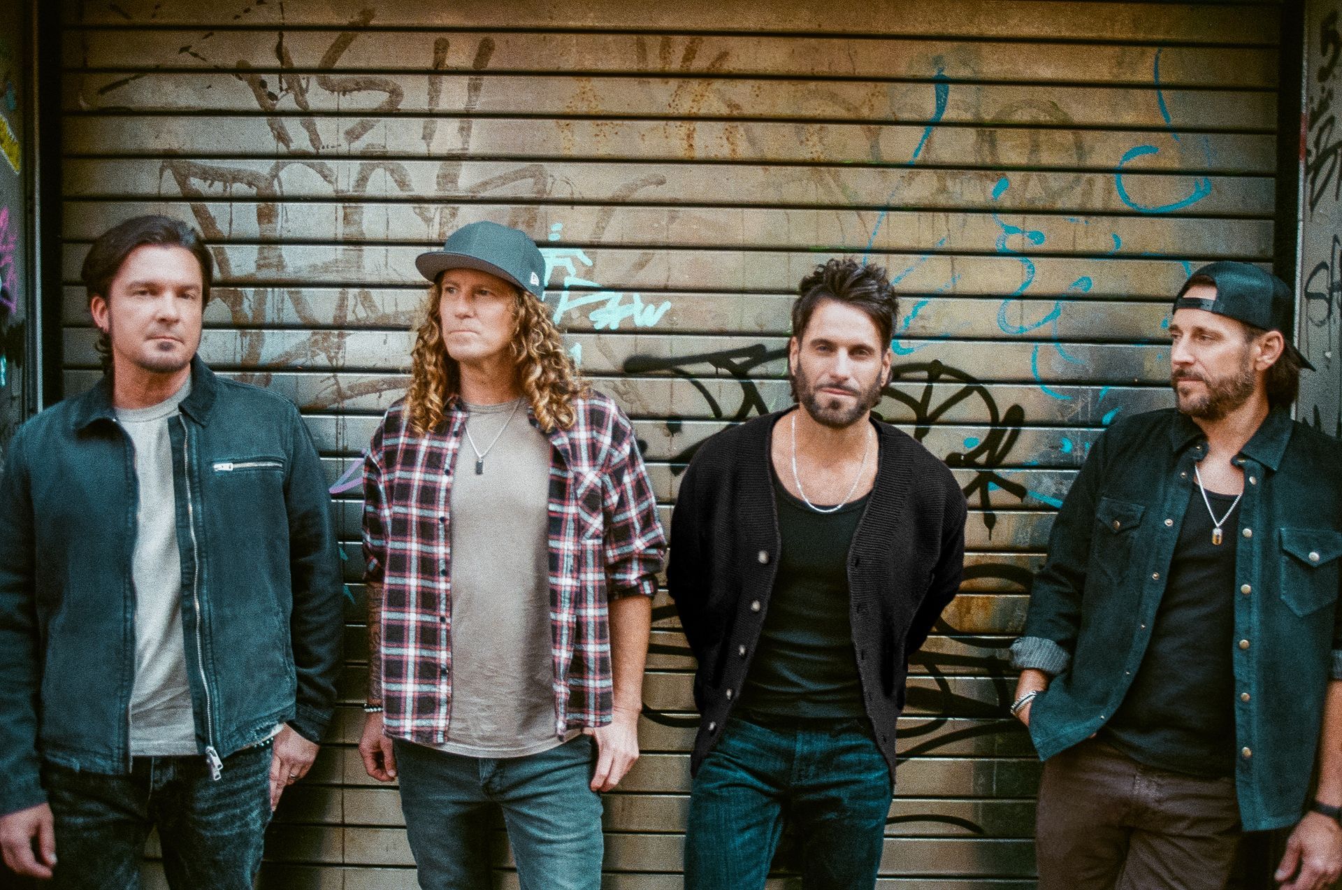 Four men posing in front of a graffiti-covered metal door. They wear casual clothes and look at the camera.