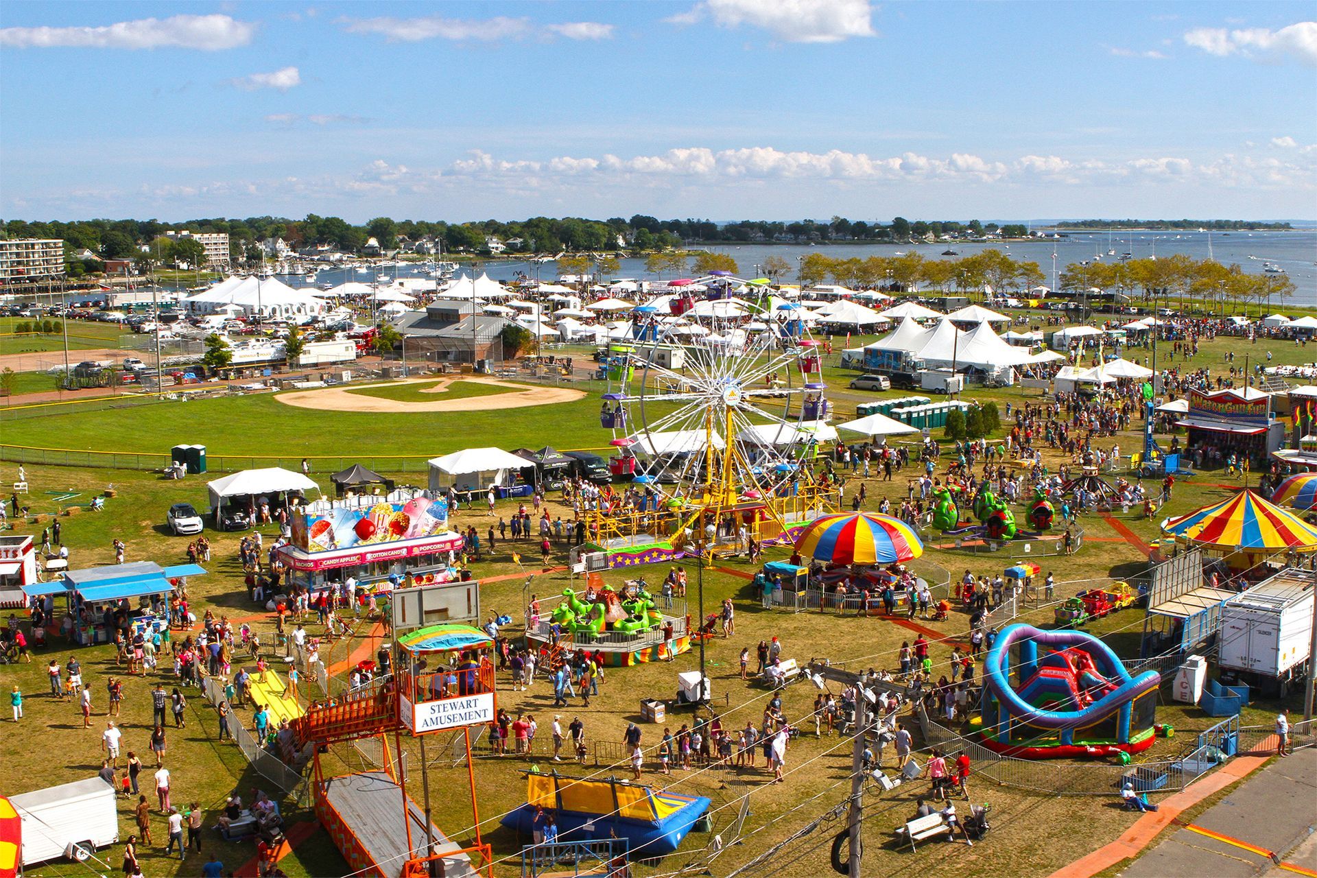 Fair on a grassy field with rides, tents, and many people; water in the background.