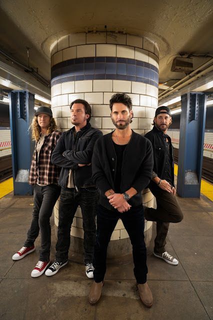 Four-piece band posing in a subway station: Casual attire, serious expressions. Brick column and blue tile backdrop.