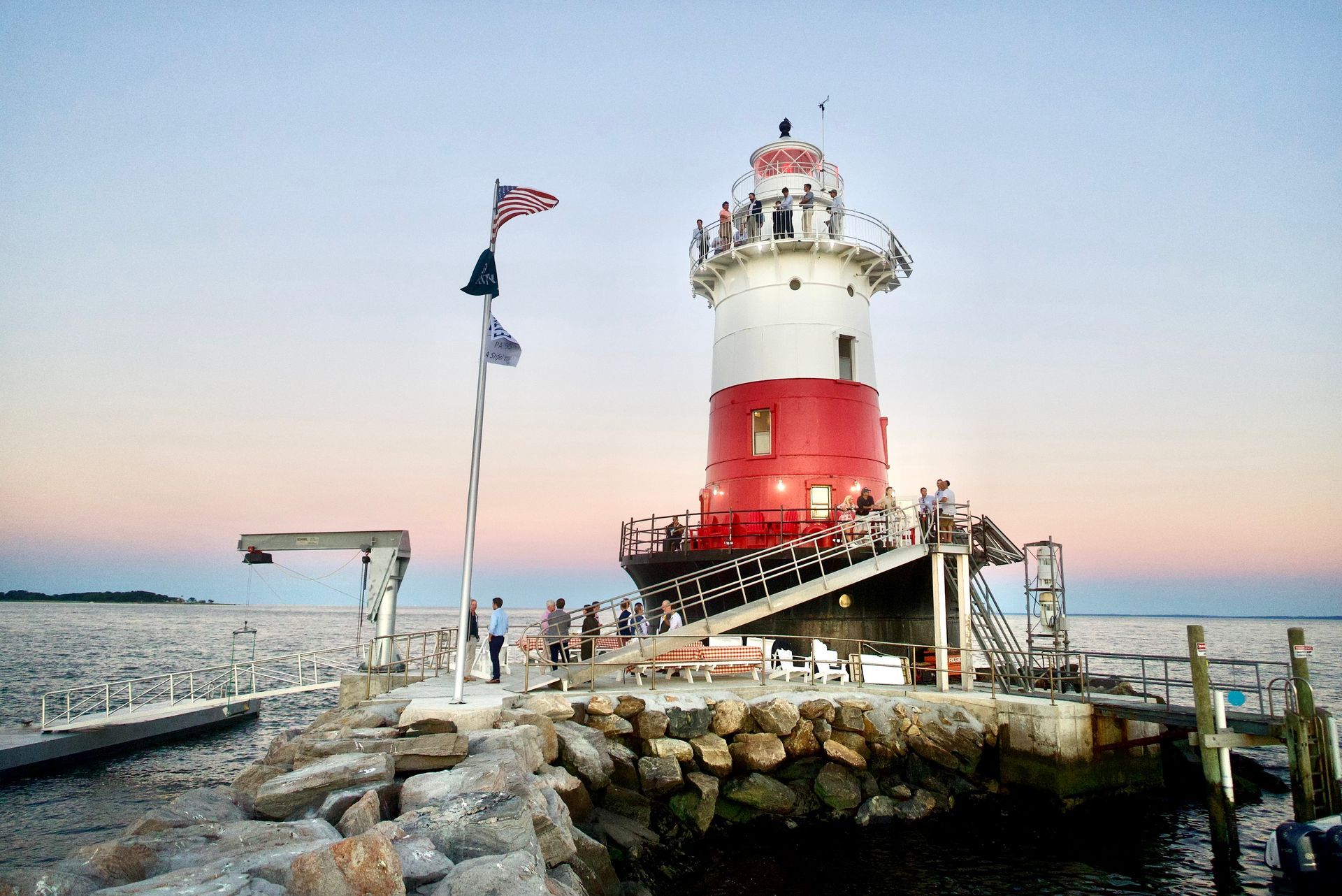 Lighthouse with red and white paint, sits on rocks by the water with people gathered around.