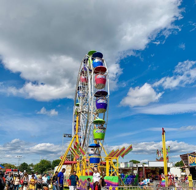 Ferris wheel at an outdoor fair against a cloudy blue sky. People milling around below.