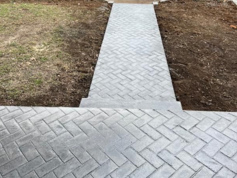 Concrete walkway with herringbone pavers leading through a dirt and grass yard