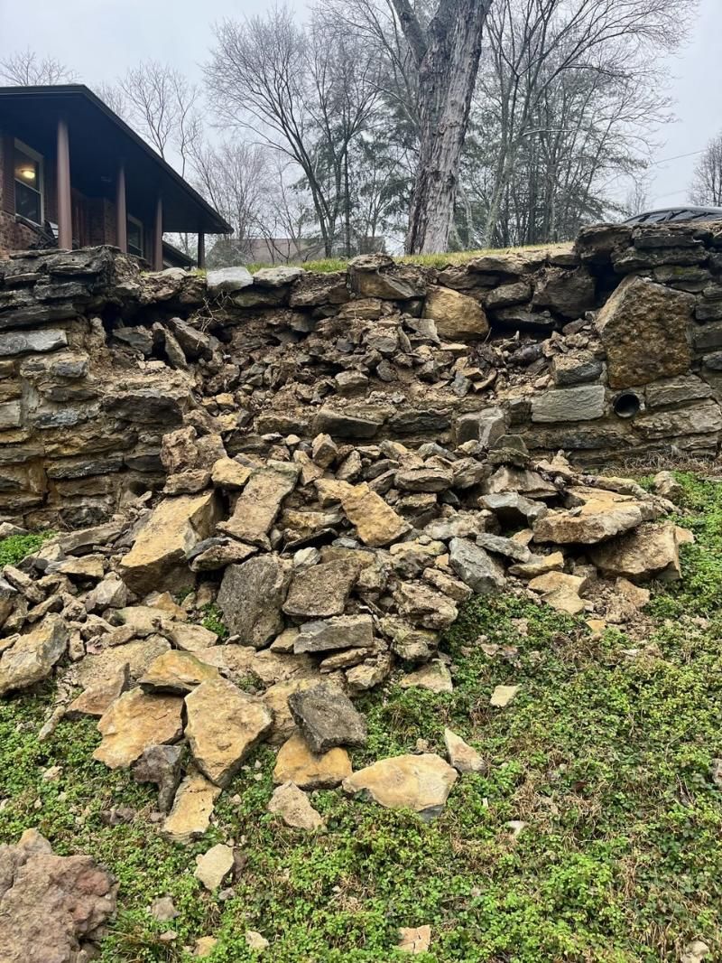 Collapsed stone retaining wall with scattered rocks beneath a house in a grassy yard