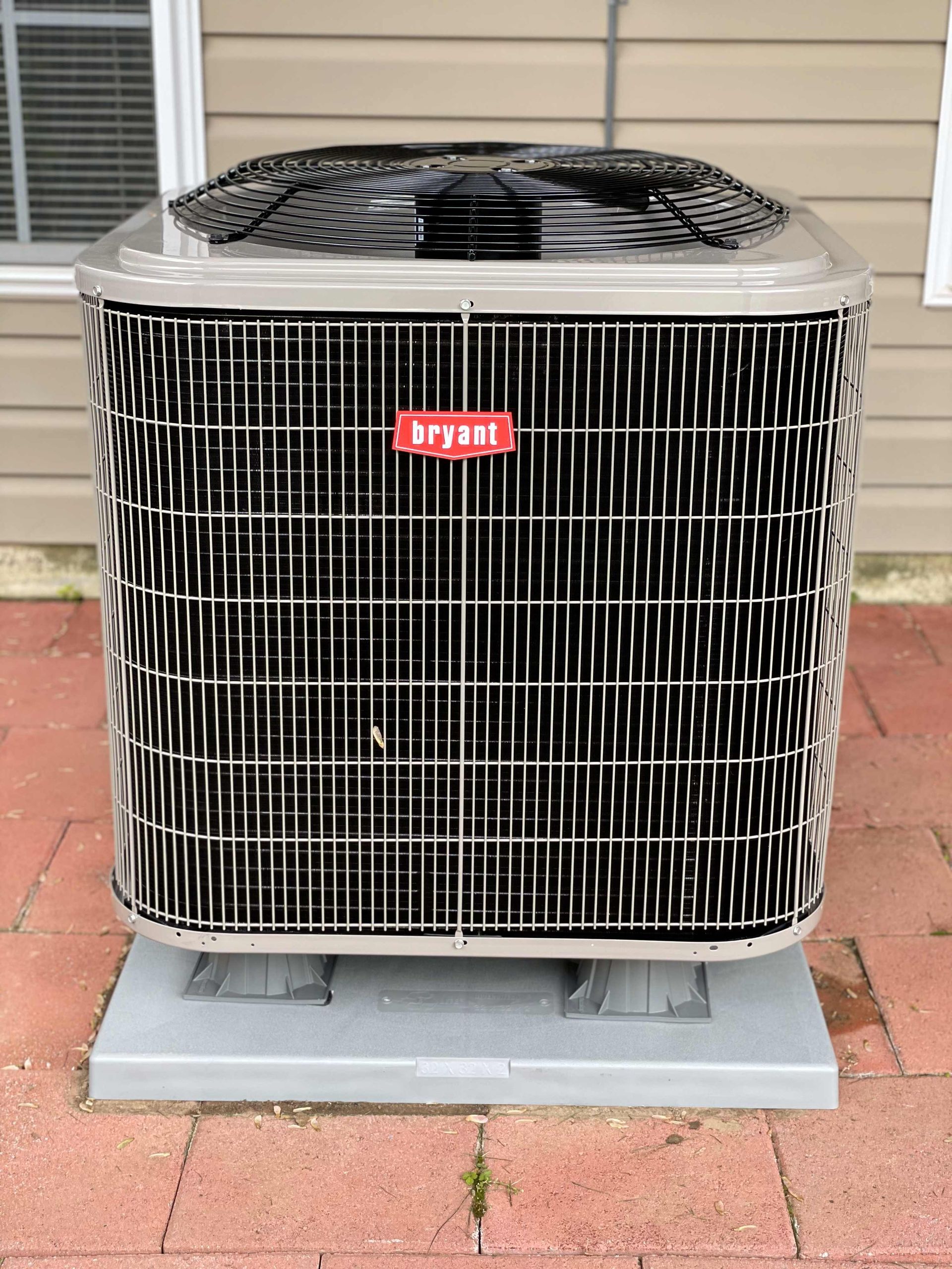 A bryant air conditioner is sitting on a brick sidewalk in front of a house.