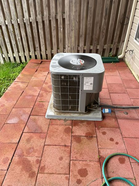 An air conditioner is sitting on a brick patio next to a hose.