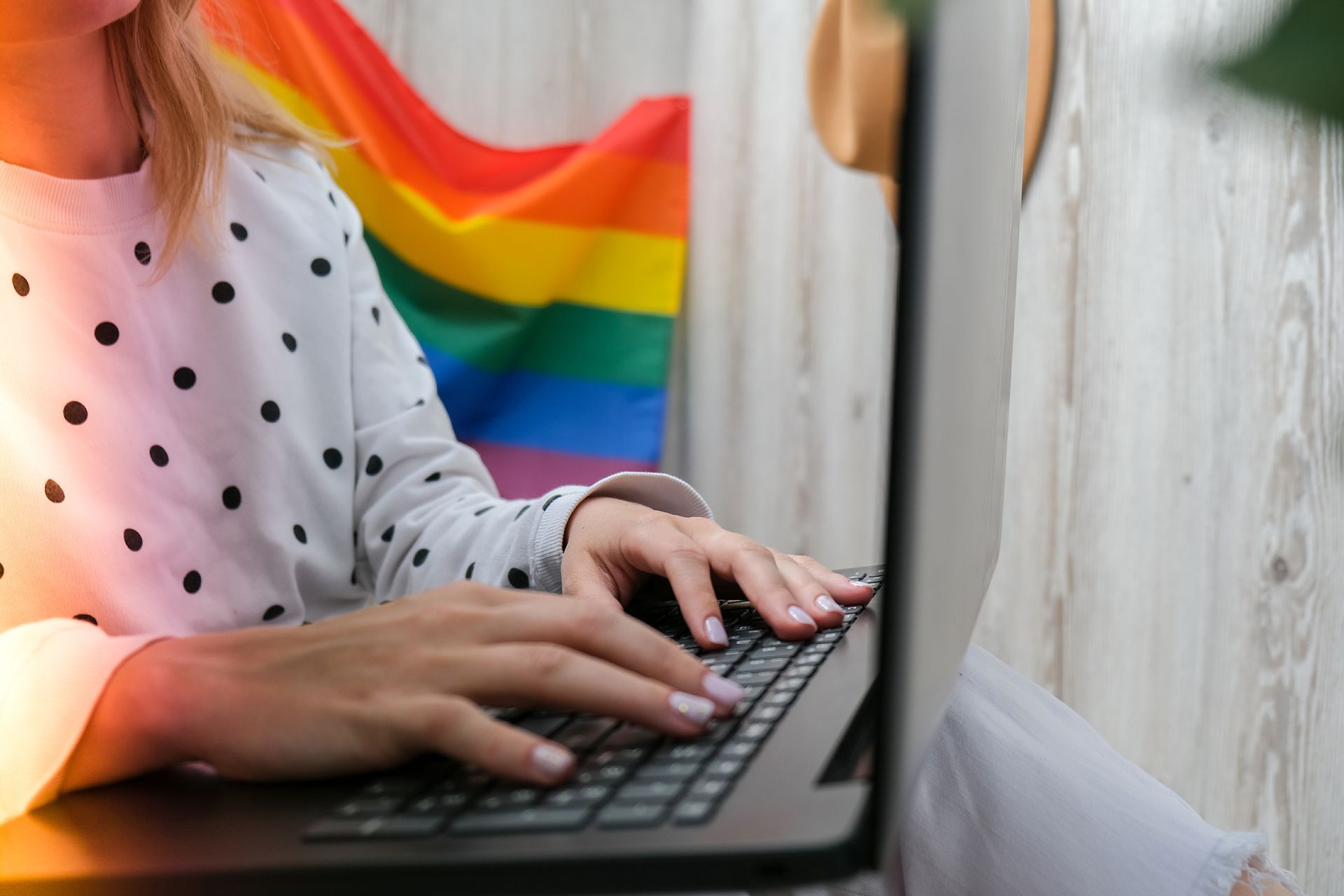 A woman is typing on a laptop computer in front of a rainbow flag.