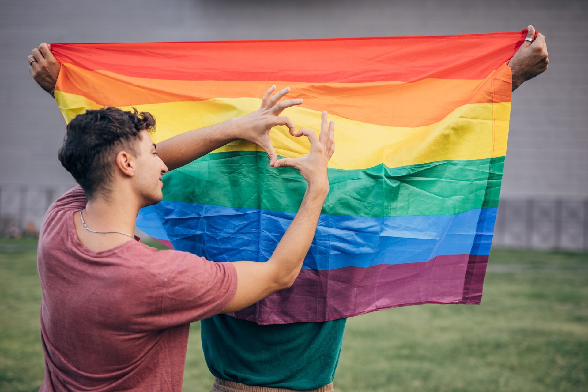 Two men are holding a rainbow flag and making a heart with their hands. Two men are holding a rainbow flag and making a heart with their hands.