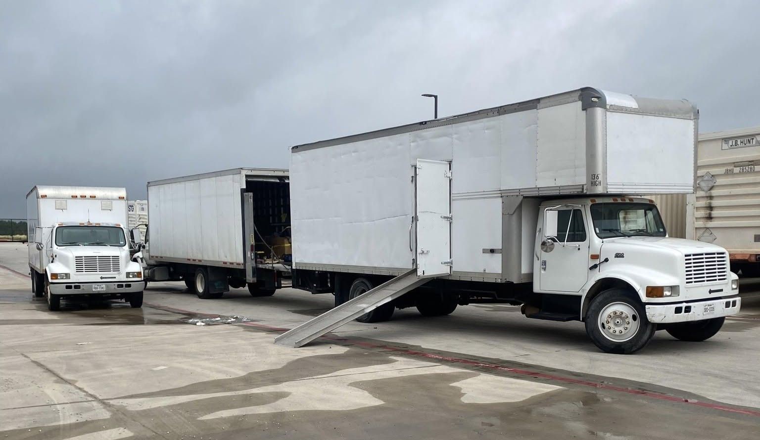 Three white box trucks parked in an industrial lot, one with a ramp extended.
