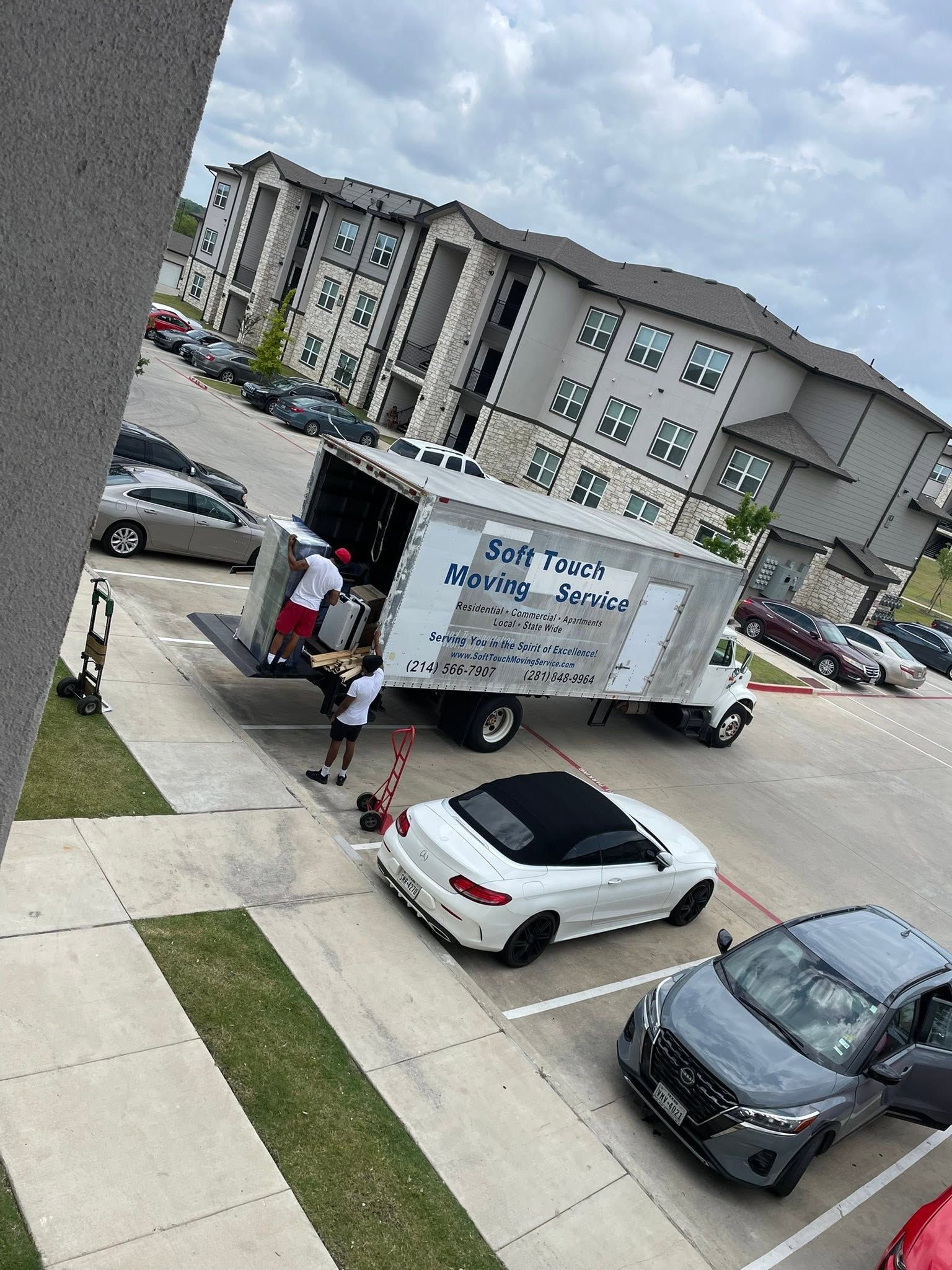 Moving truck outside an apartment complex, with people loading items beside parked cars.