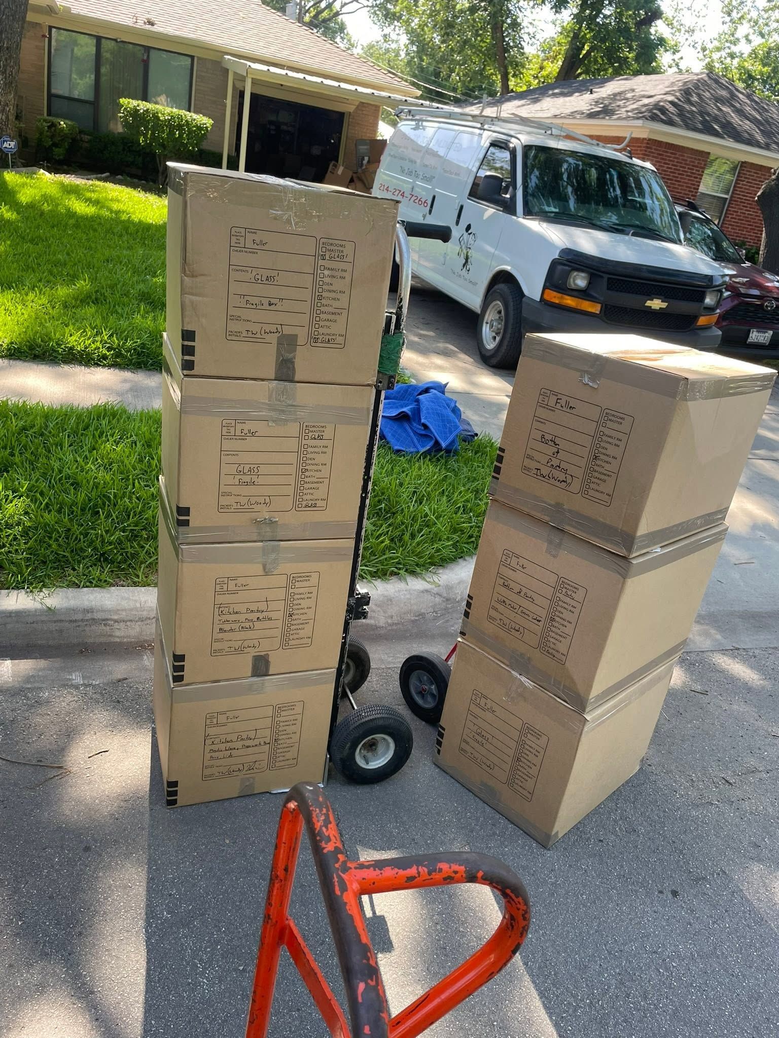 Stacks of cardboard boxes on a driveway beside a white van and red hand truck in a suburban yard.