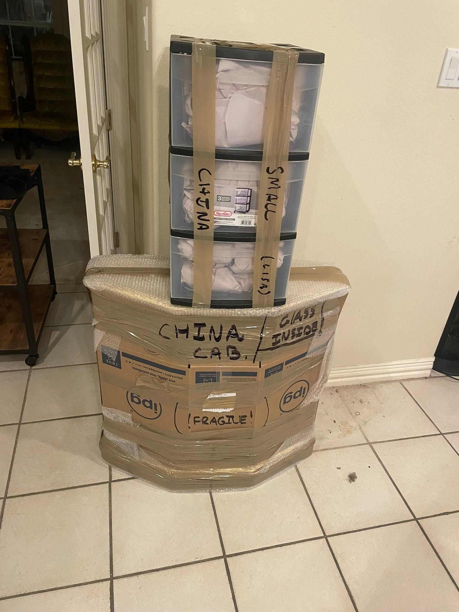 A taped cardboard package with a small framed object on top, sitting on a tiled floor against a wall.
