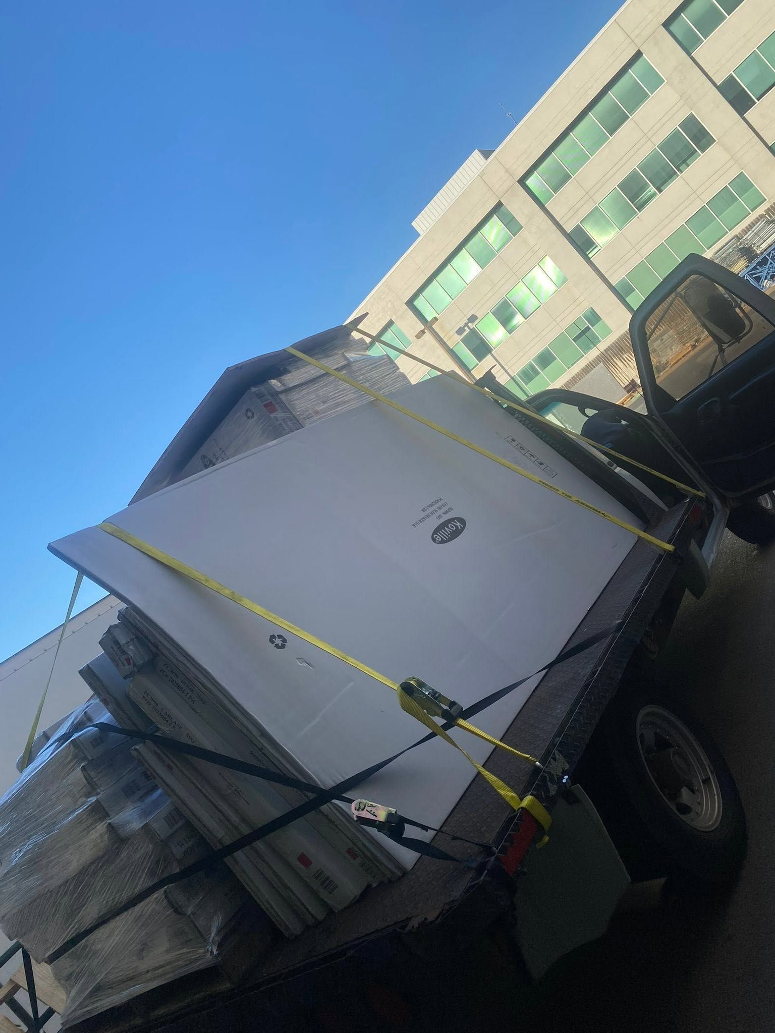 Overturned white truck beside a building, with black straps securing its load under a blue sky.