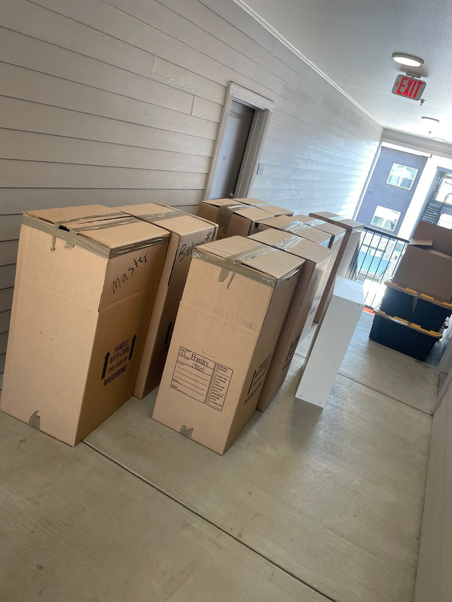 Cardboard boxes stacked in a hallway near a glass door with an exit sign.