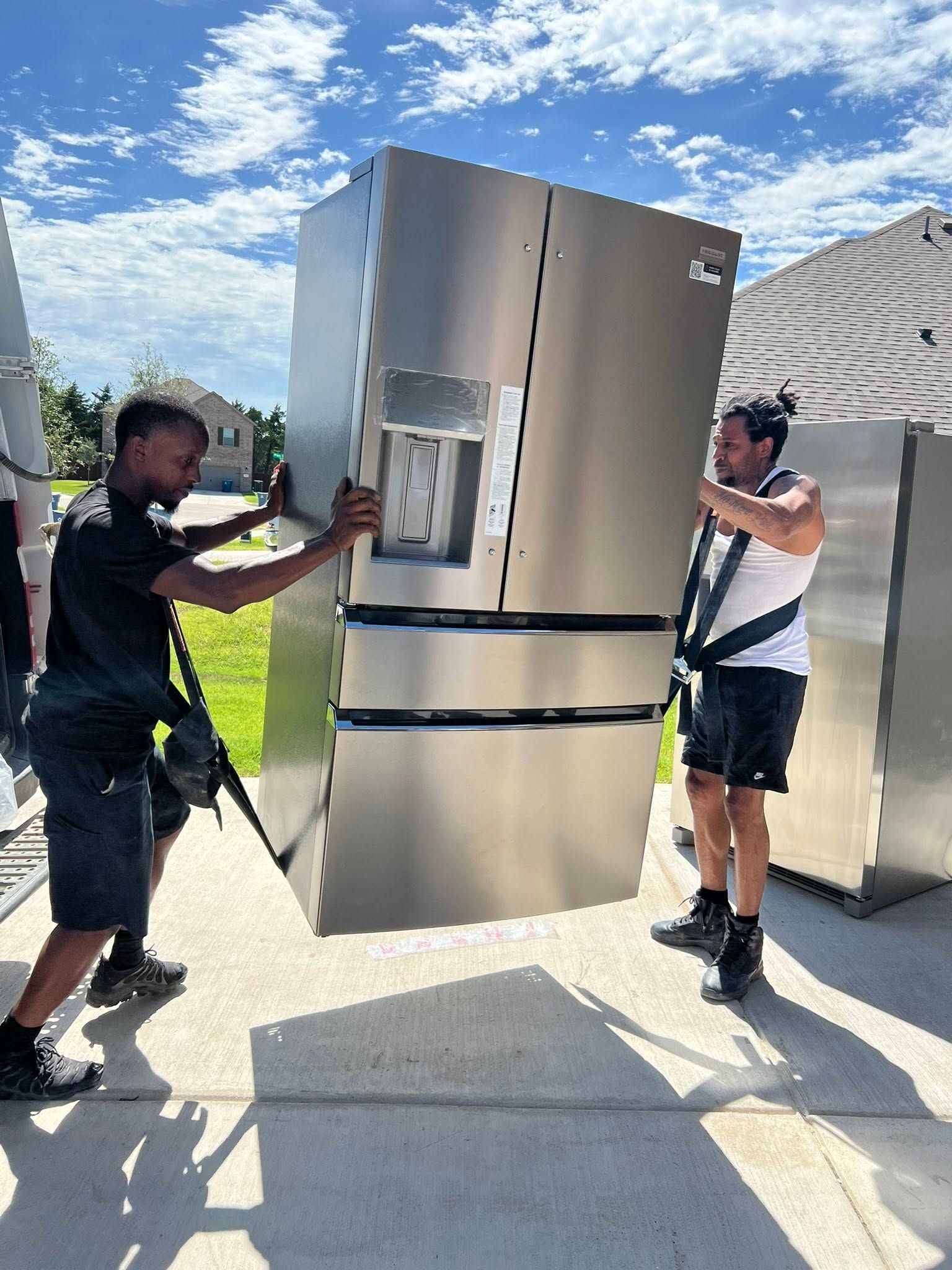Two men moving a stainless steel refrigerator outdoors on a sunny driveway.