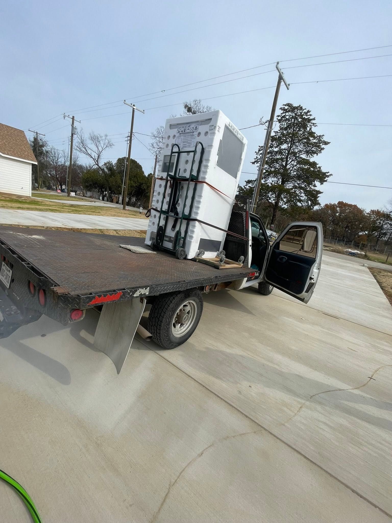 Flatbed tow truck carrying a white refrigerator on a roadside, with the truck door open.