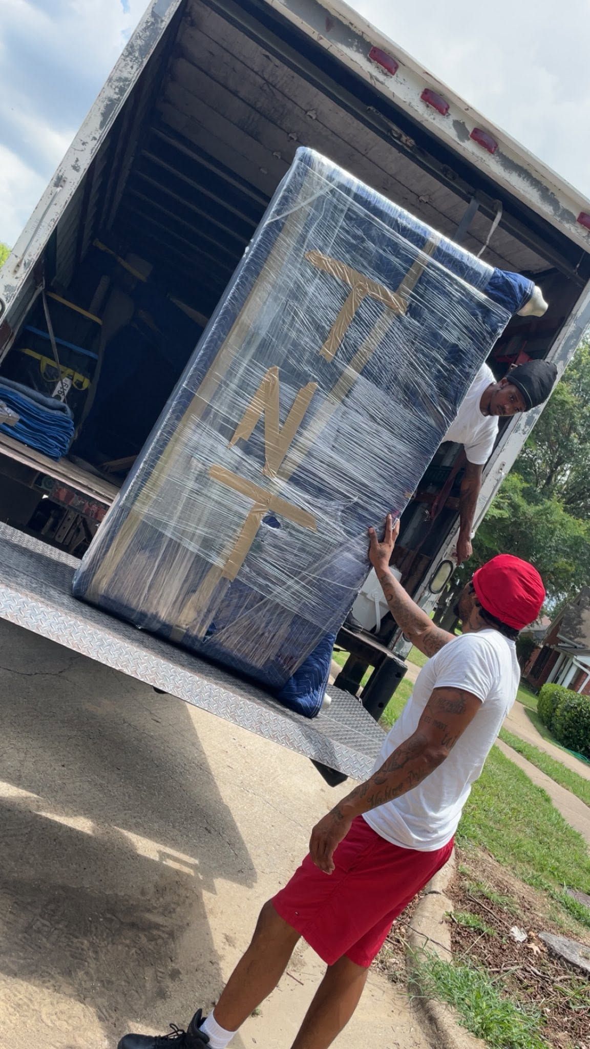 Person in red shorts loading a wrapped mattress into a truck from a driveway.