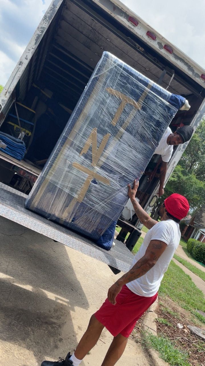 Man unloading a wrapped mattress from a truck on a sidewalk, wearing a red cap, white shirt, and red shorts.