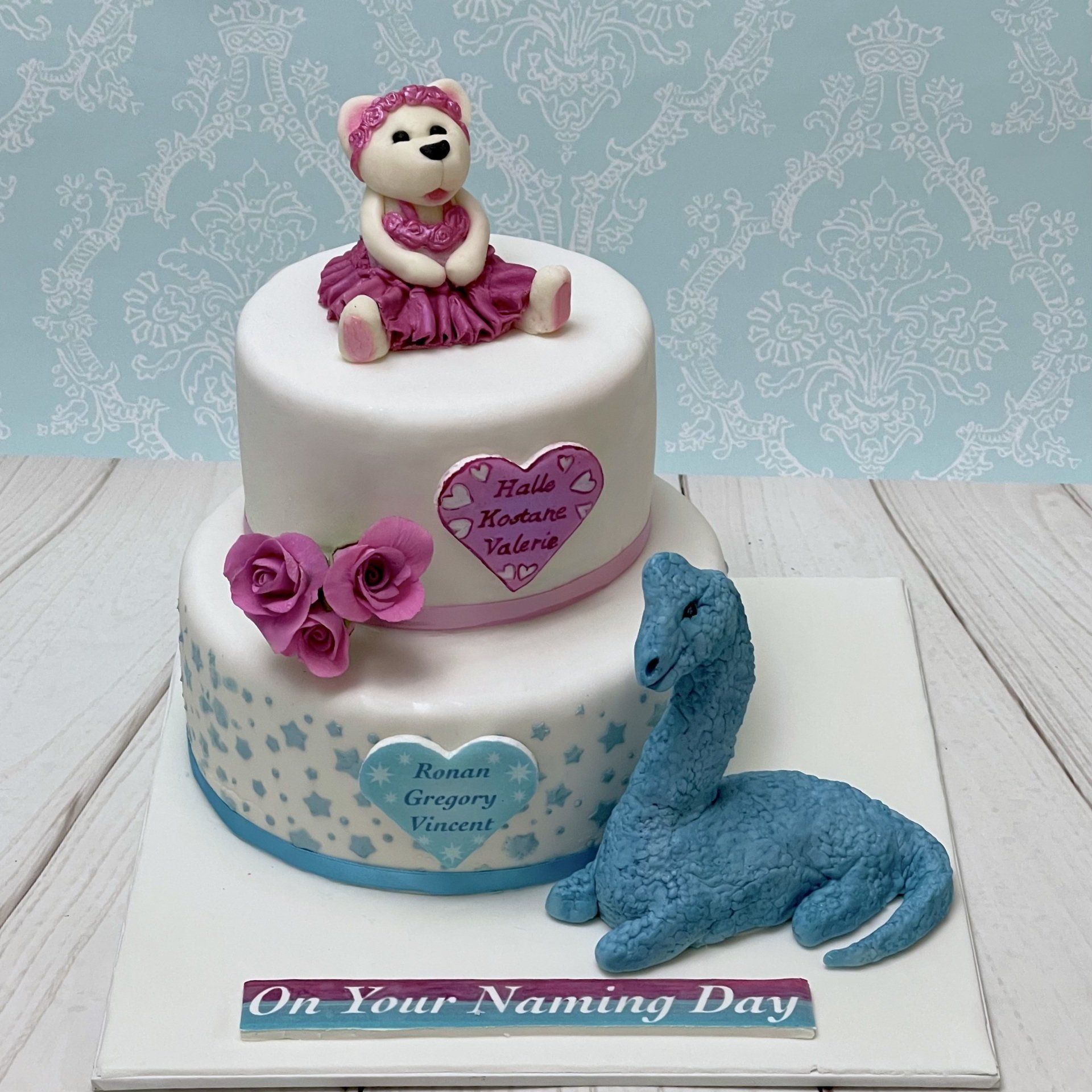 Two Tier Naming Day Cake. Top tier White fondant with a cute fondant ballerina bear, and pink sugar roses Bottom tier with blue stars and a fondant dinosaur on the board, both tiers have a heart plaque also with the children's names.