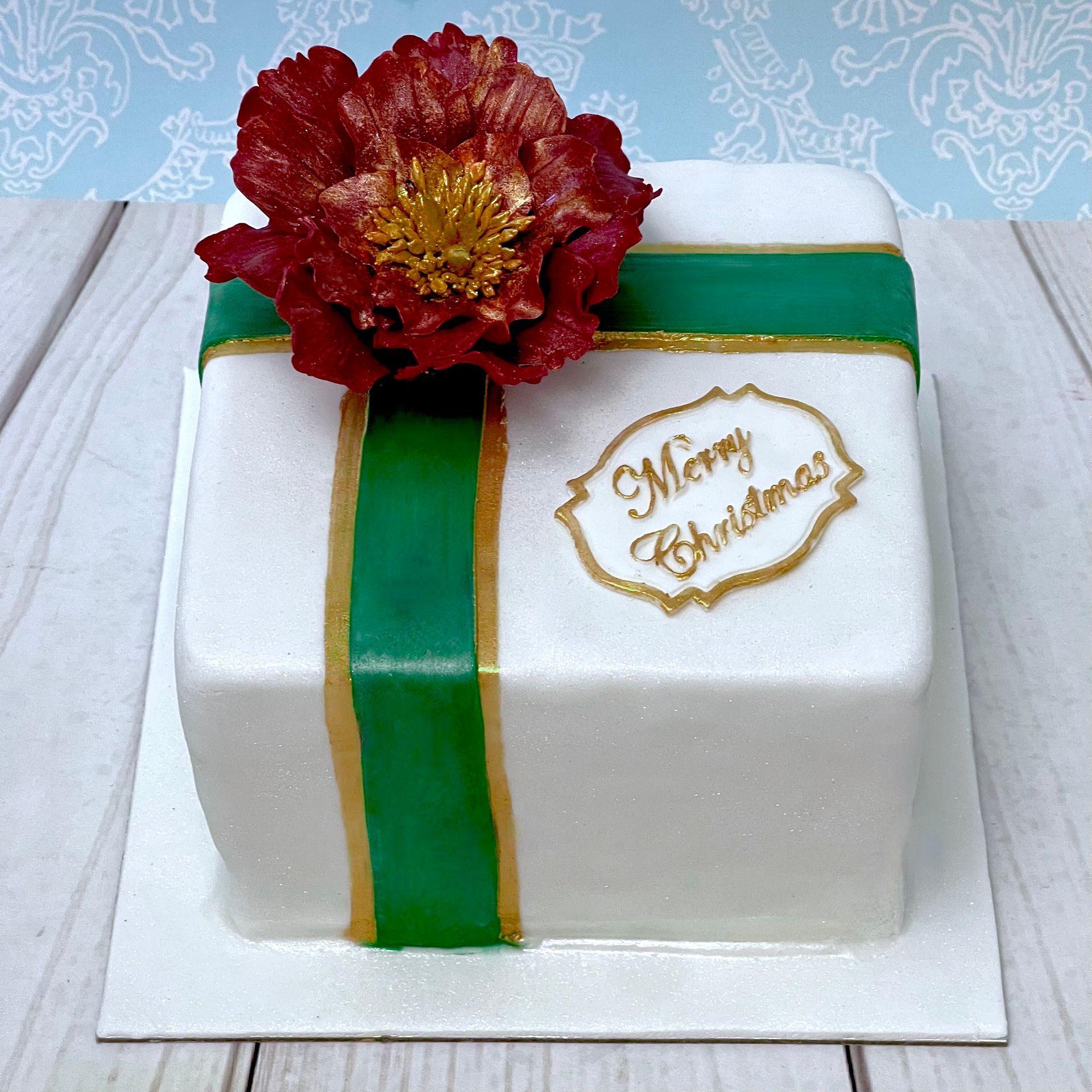 Christmas cake covered in fondant decorated to look like a Christmas present with green and gold fondant ribbon a Merry Christmas fondant tag and a red and gold sugar flower. 