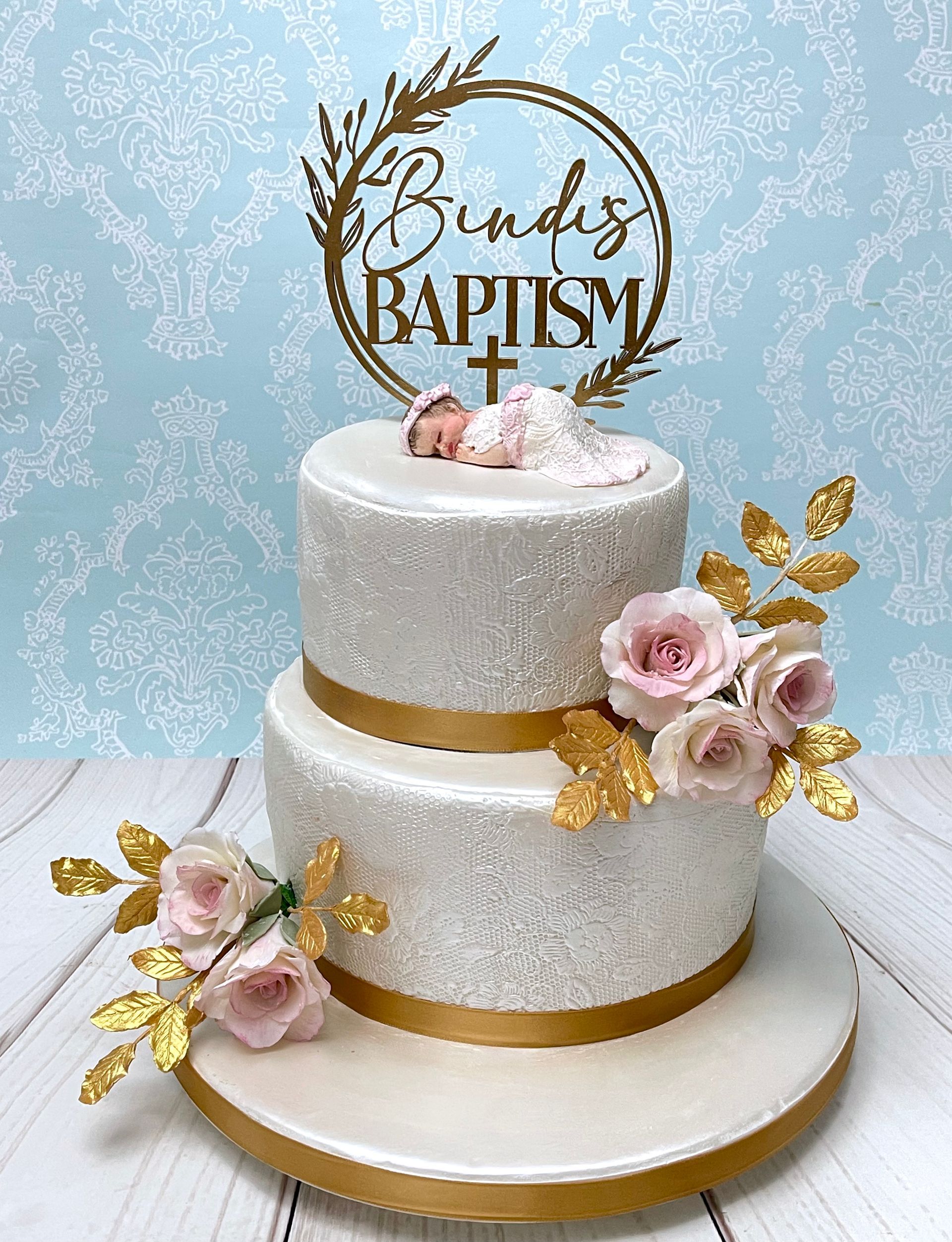 Elegant Two Tier Baptism cake Port Macquarie. With lace effect around sides, a hand sculpted fondant baby topper in a white fondant lace gown, A Personalised  baptism topper, white and soft pink sugar roses and gold sugar leaves 