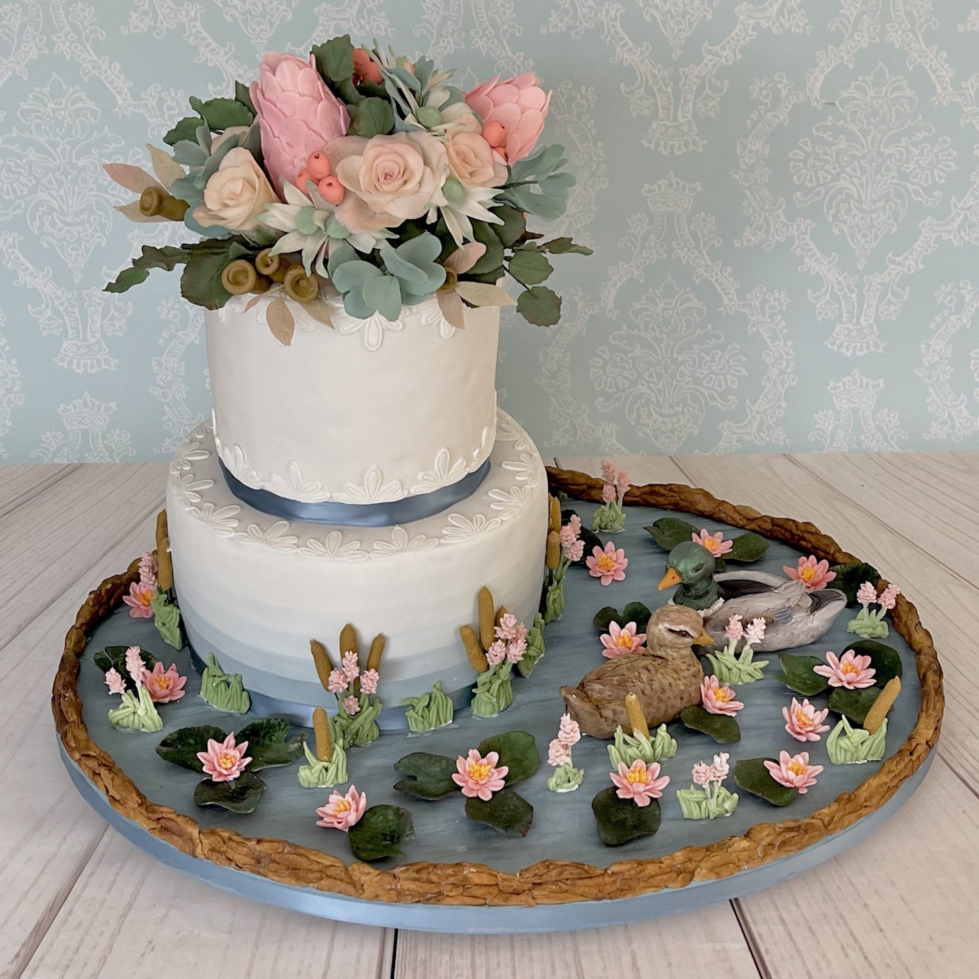 Gorgeous Two tier Duck pond wedding cake. The top tier of the wedding cake has a beautiful arrangement of sugar roses and sugar Australian native flowers, the cake is surrounded by a beautiful sugar waterlily pond with two very cute sugar ducks. Created For Celebrity Dr Harry Cooper and His Wife Sue.