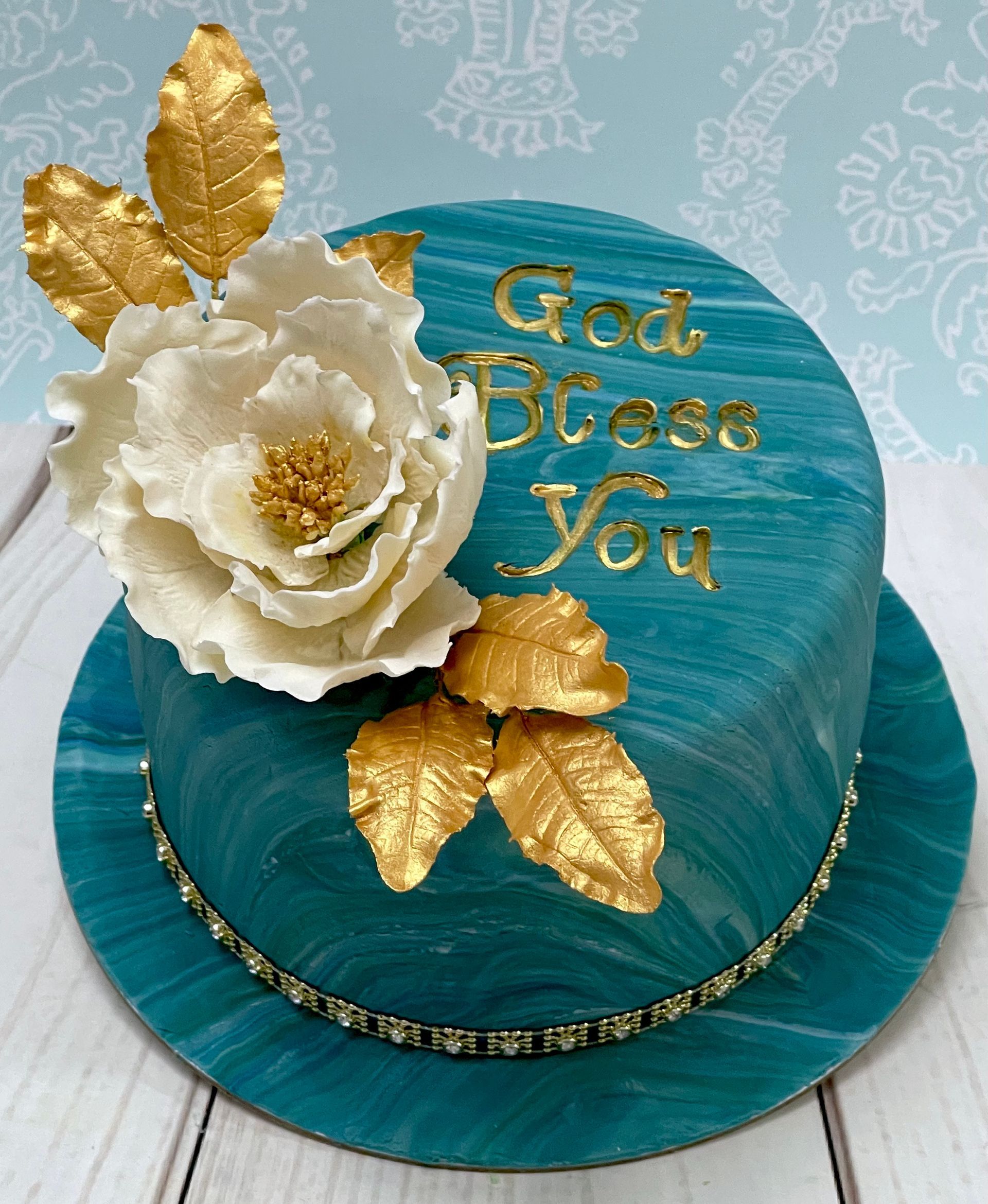Blue and teal marbled fondant cake with hand painted God Bless You, White Sugar Open Rose and Gold sugar Leaves.