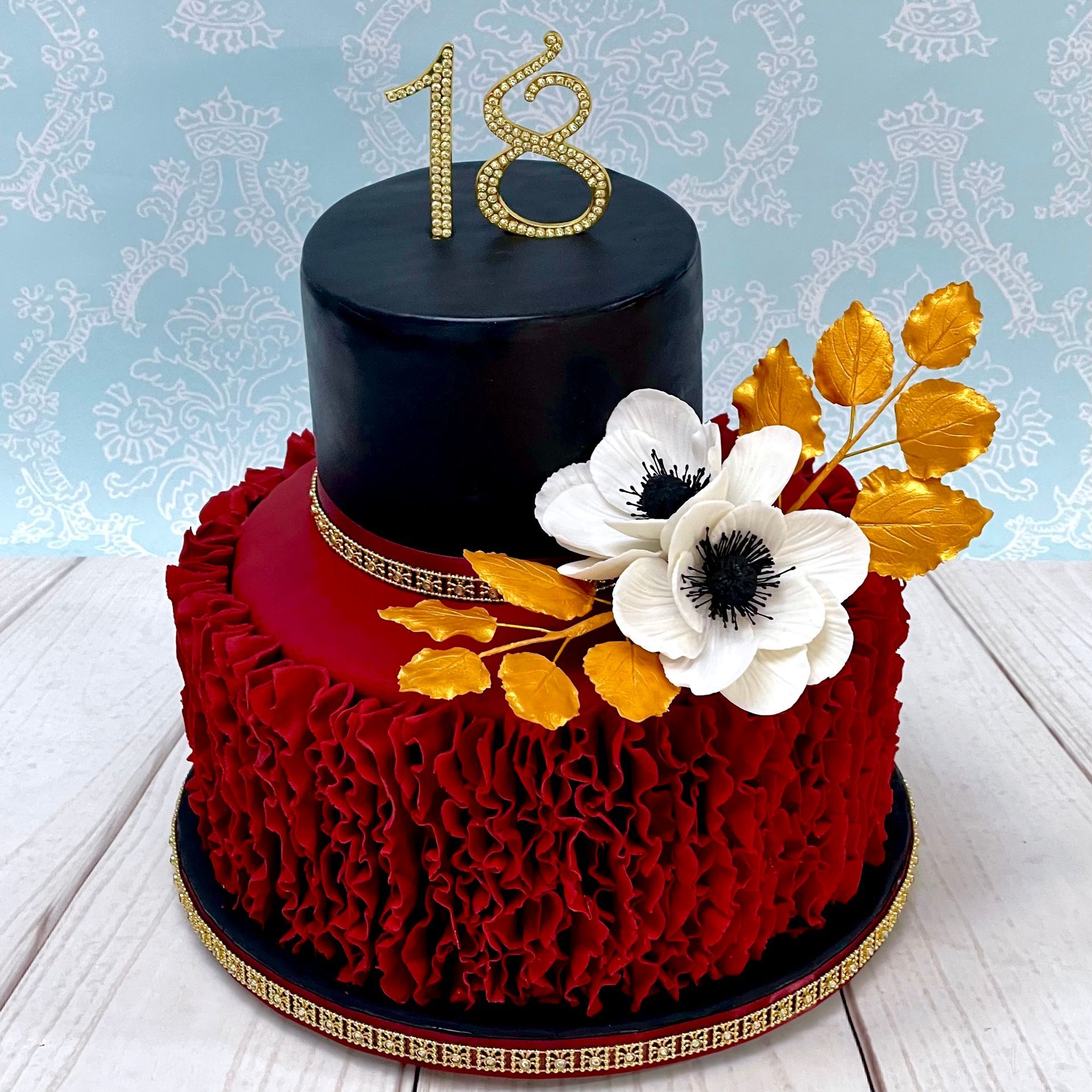 Glamerous 18th Birthday Cake, 
Base tier red ruffles, 
top tier black shimmer, with White sugar aneomone flowers, gold sugar leaves, and a gold diamante 18 and trim.