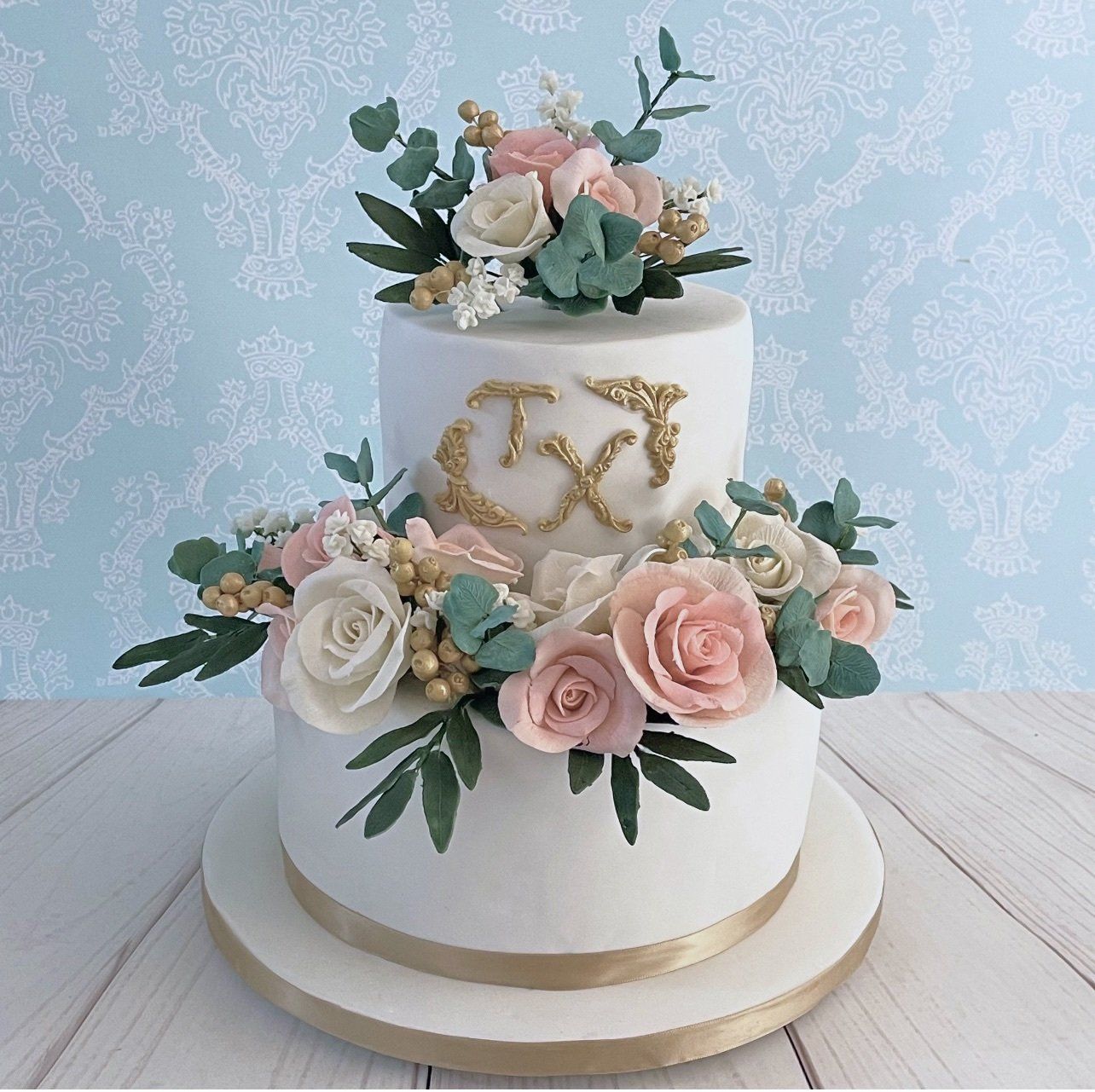 Two tier wedding cake. White fondant Champagne gold ribbon around the base of each tier and board, Champagne gold filagree Initals on front of top tier and Sugar flower arrangements consisting of sodt pint and white roses and champagne gold sugar berries, sugar babies breath, and sugar eucalyptus leaves