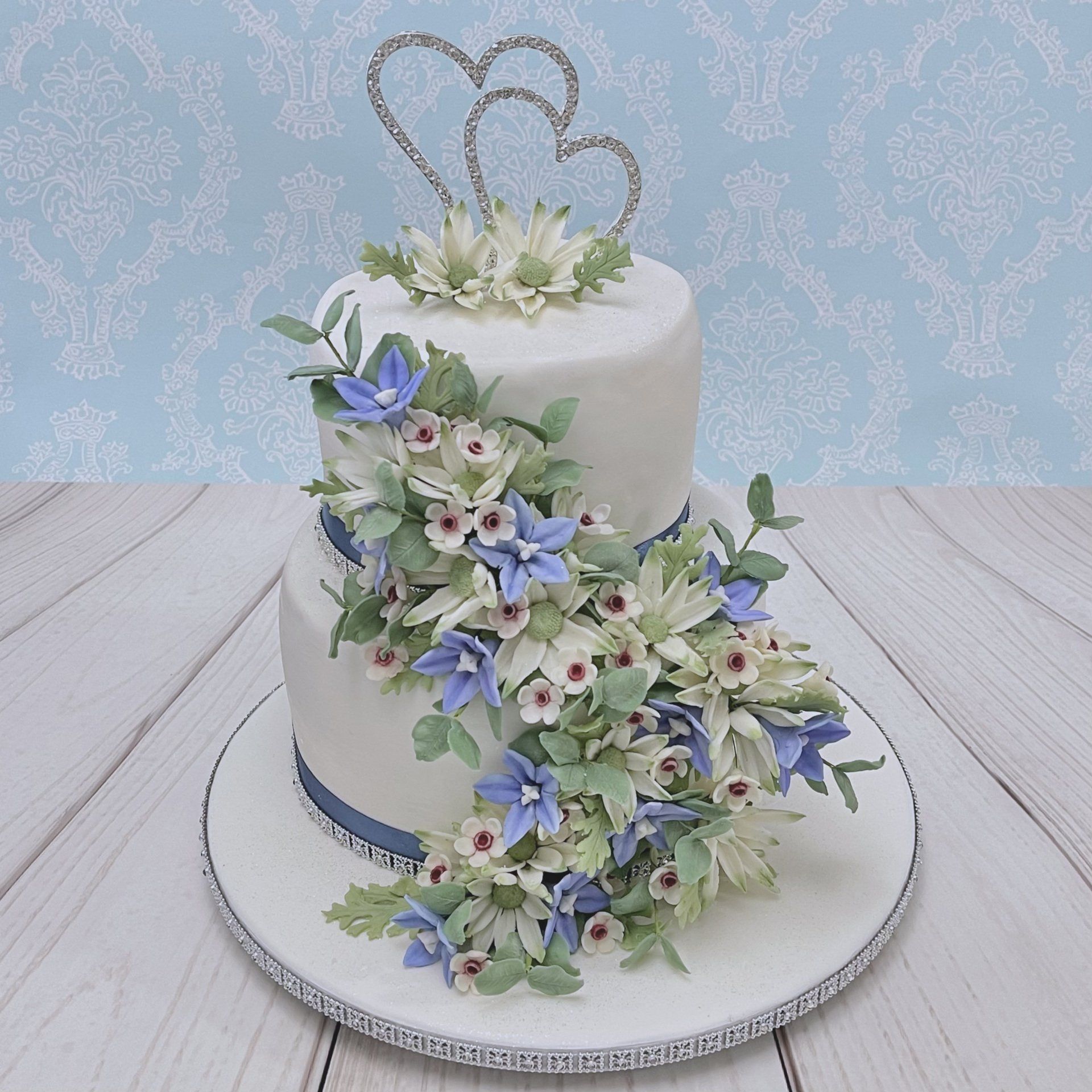 Two tier Wedding cake. White fondant with dusty blue ribbon and diamanté trim and a cascade of Ausralian Native sugar flowers including Flannel Flowers, Australian blue bells, geraldton wax, and eucalyptus leaves. Finished with a Diamanté double heart topper.