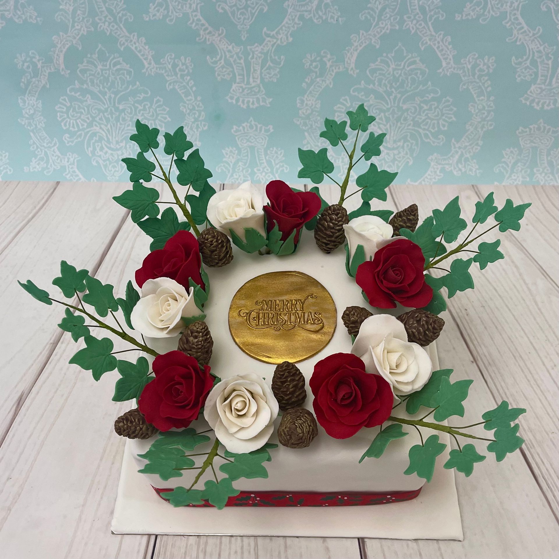Christmas Wreath Cake With Sugar Roses, Pine Cones and ivy Leaves with a gold plaque that says Merry Christmas.