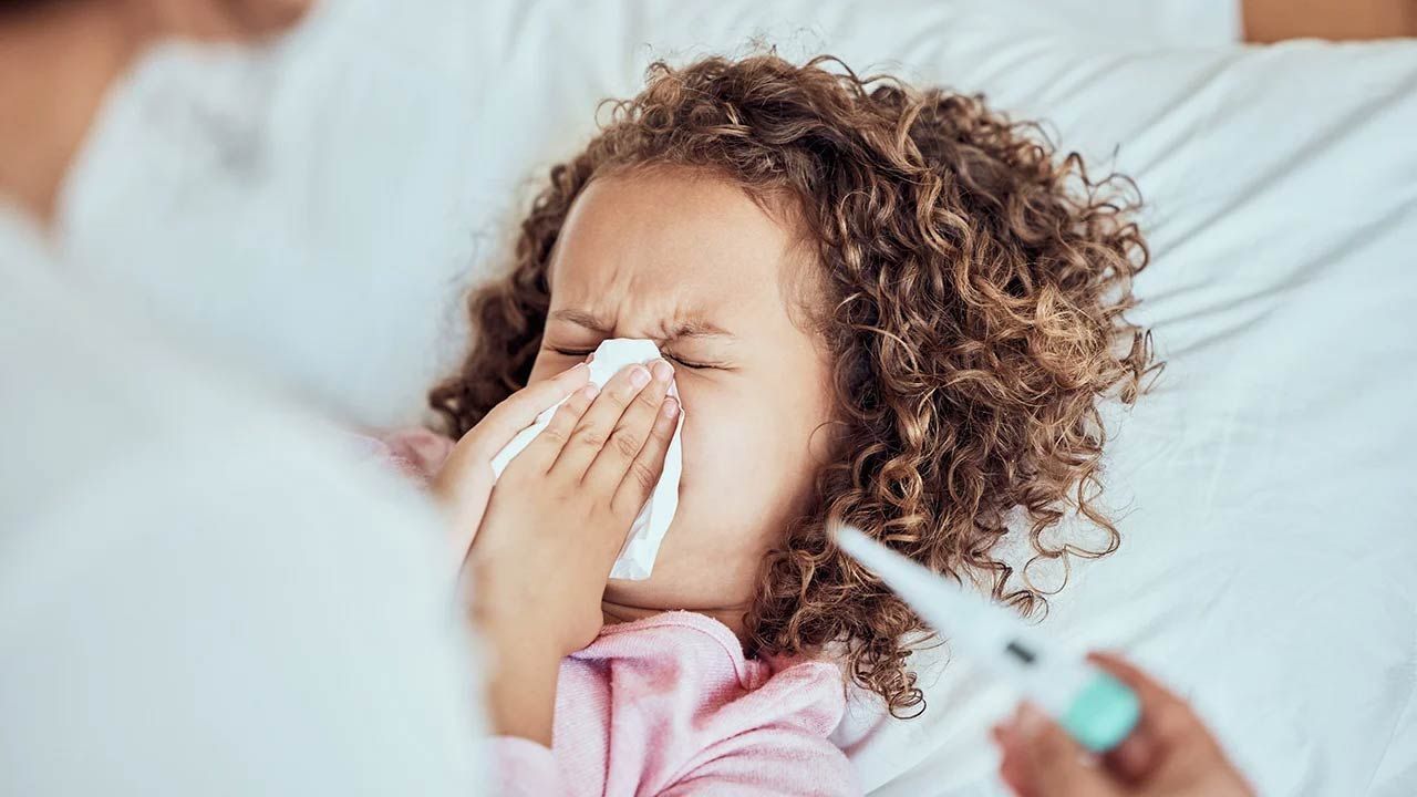 Child sneezing into a tissue while a hand holds a thermometer nearby