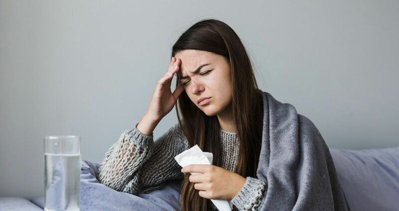 Woman wrapped in a blanket, holding a tissue and touching her forehead beside a glass of water