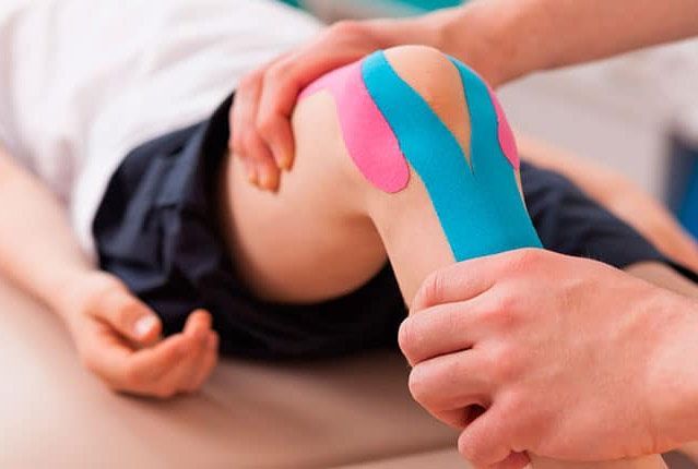 Hands applying blue and pink tape to a bent knee on a treatment table