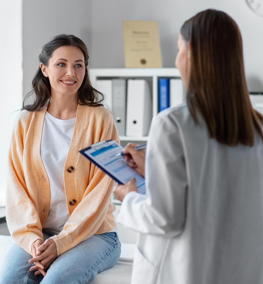 Two women in a clinic, one smiling while speaking with a doctor holding a clipboard.