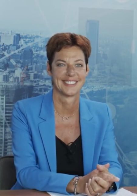Lachende vrouw in een blauwe blazer zit voor een stadsgezicht op de achtergrond.