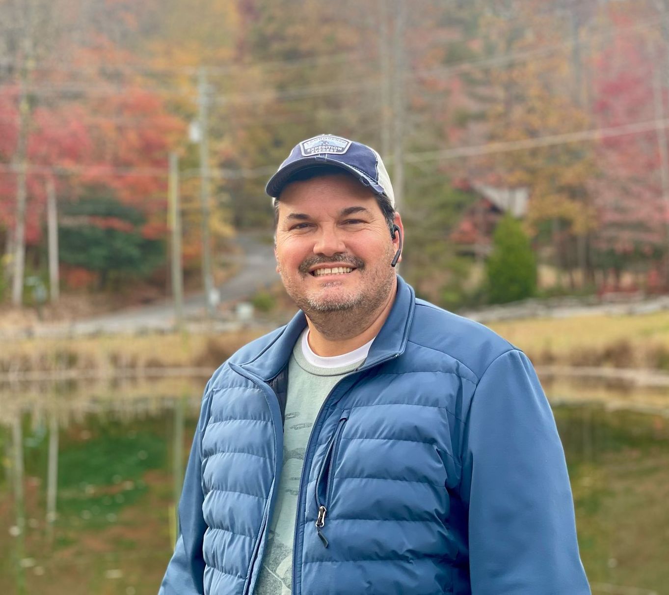 Man smiling, wearing blue jacket and hat, standing near a pond in front of fall foliage.