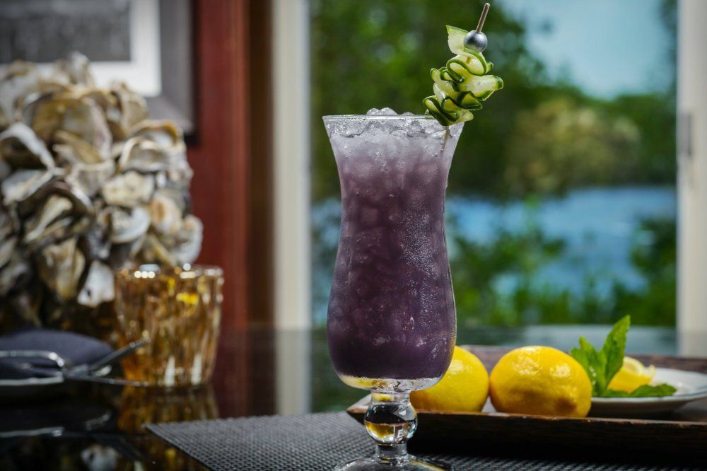 Purple cocktail with mint garnish on a table, beside lemons and a blurred floral arrangement and lake view