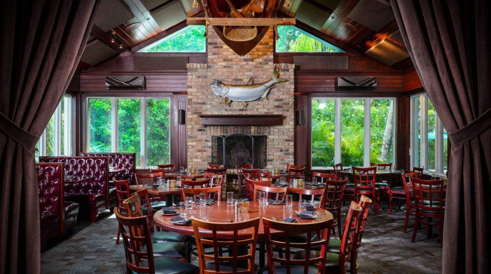 Rustic lodge dining room with wooden tables, chairs, large windows, and a stone fireplace at the center