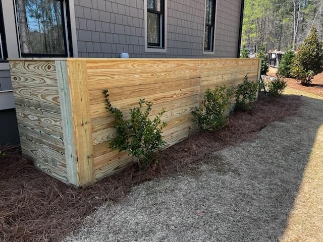 Wooden fence covering an outdoor unit, with shrubs planted along its base. Gray house in background.