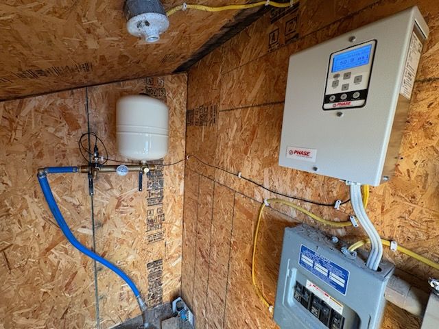 Interior of a wooden shed with water tank, electrical panel, pump control, and wiring.