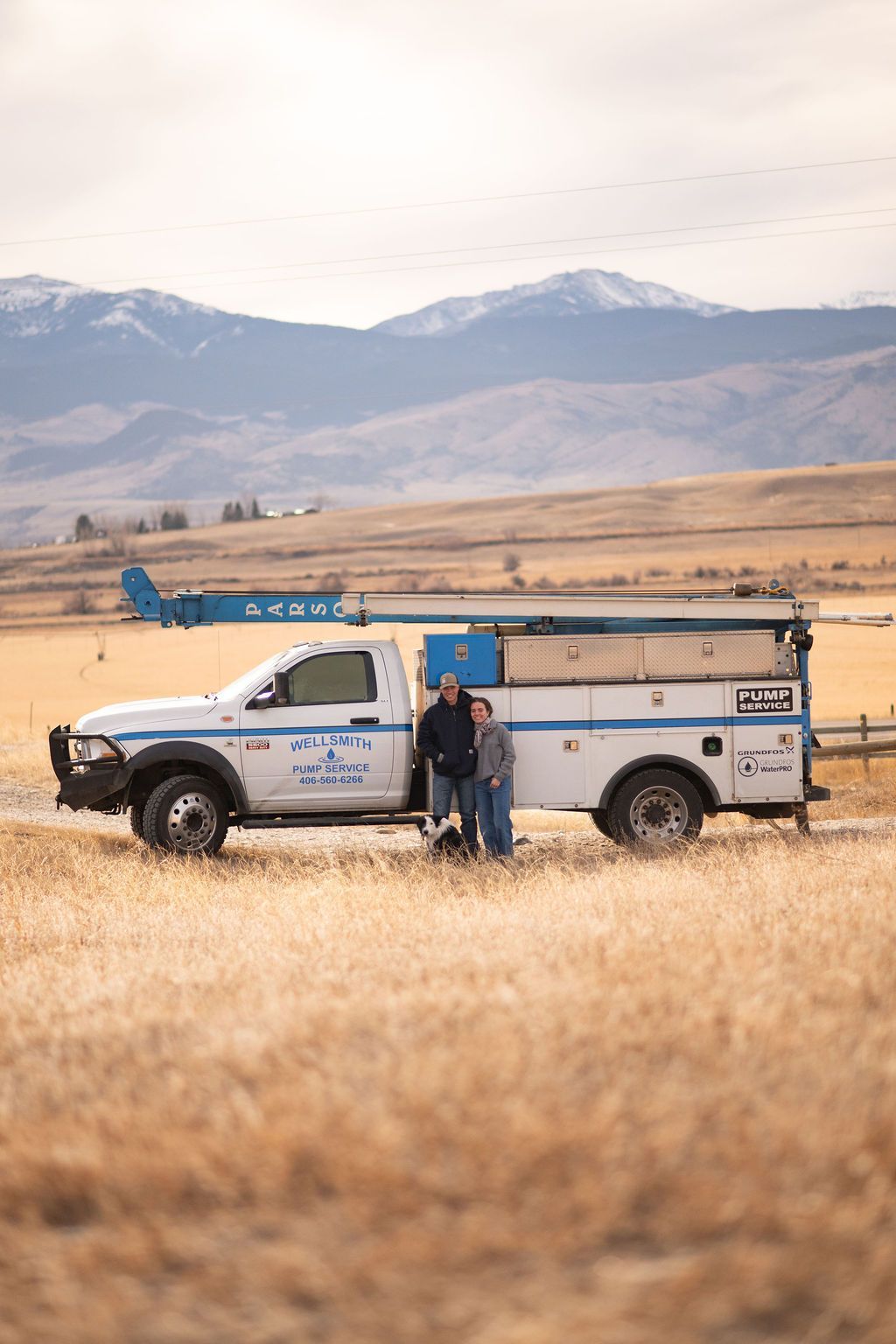 Two people stand by a white utility truck with a crane in a dry, grassy field against a mountain backdrop.