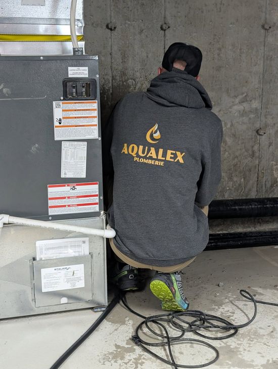 Une personne travaille sur un système de climatisation dans un sous-sol. Elle porte un sweat-shirt gris et est accroupie près de l'appareil. Un câble noir est posé au sol.