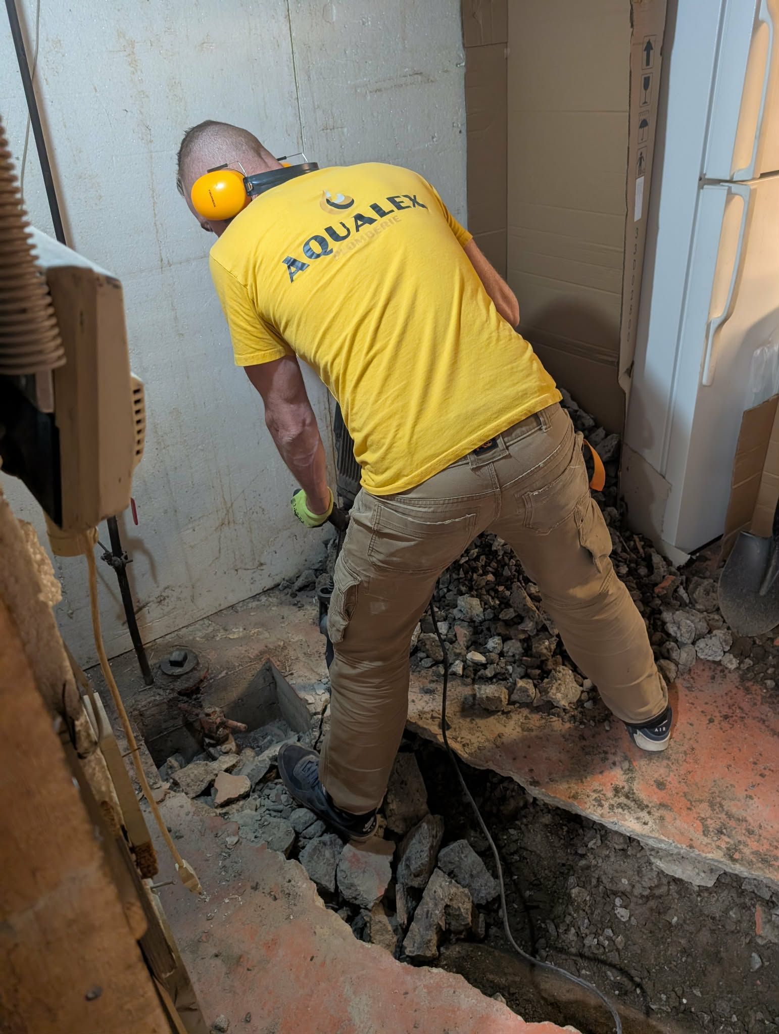 Un homme portant une chemise jaune et des protections auditives creuse une tranchée à l'intérieur d'un bâtiment à l'aide d'un marteau-piqueur.