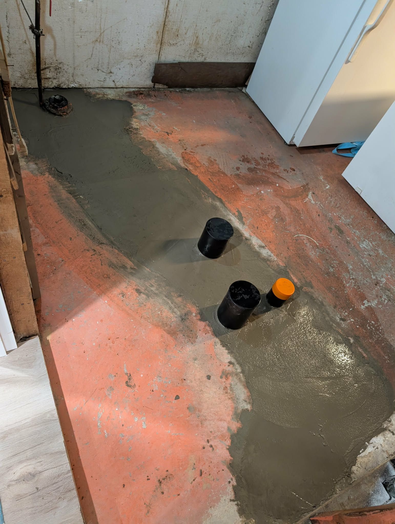 Réparation de béton au sous-sol. Tuyaux recouverts de béton gris sur sol rouge. Pompe de puisard et appareil électroménager blanc visibles.