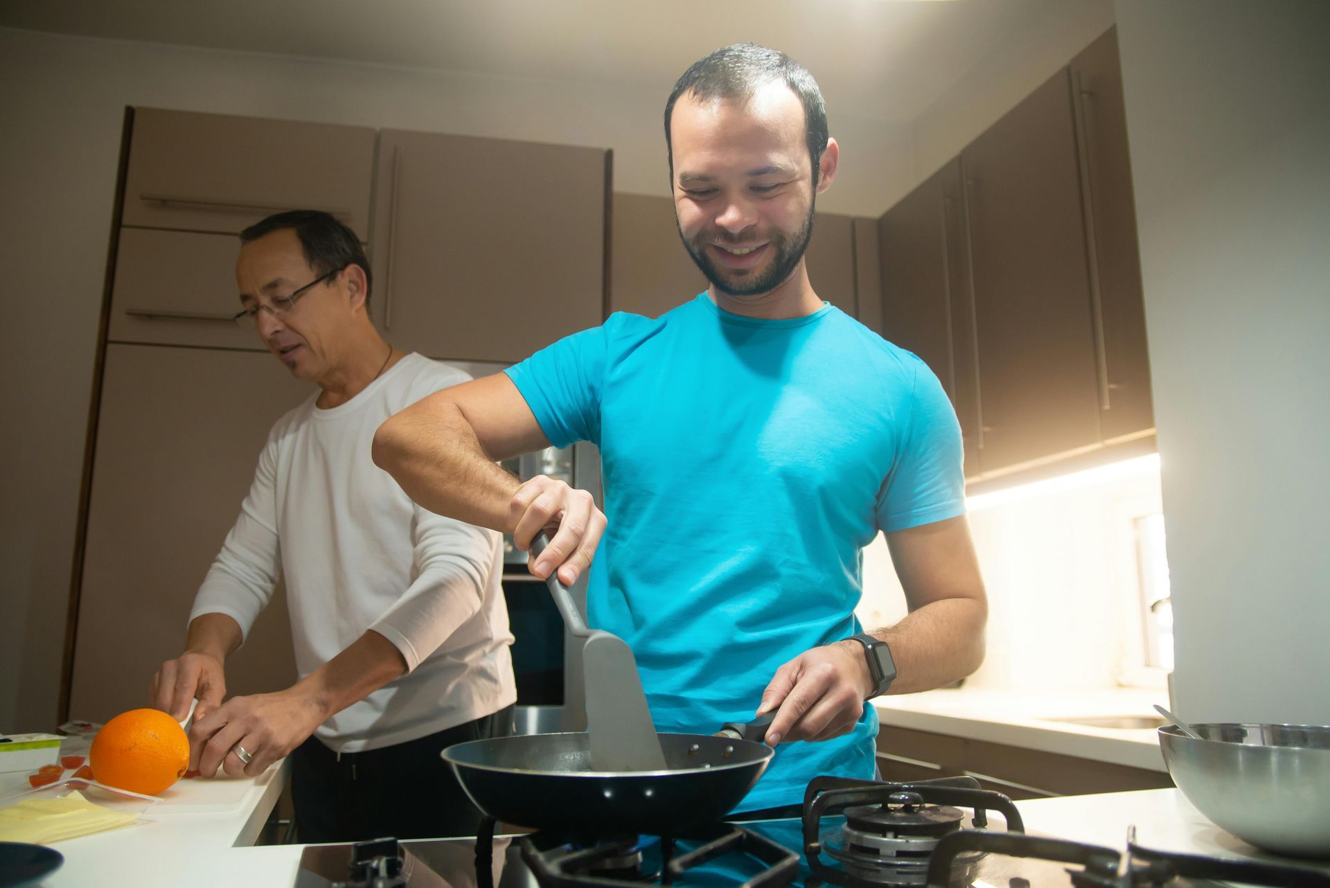 Two men are cooking in a kitchen and one is cutting an orange.